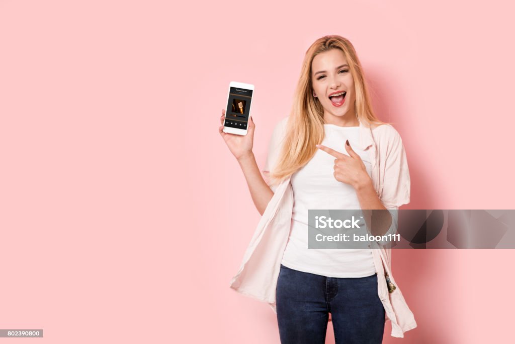 Young blonde girl is enthusiastic and points at her smartphone Using Phone Stock Photo