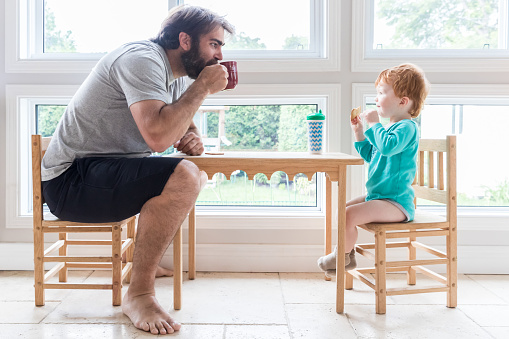 Father and Son enjoying a Sunday morning on a kid table.  The father is drinking coffee while the baby boy is drinking milk and eating a cookie.