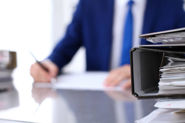 istock Binders with papers are waiting to be processed with businessman back in blur. Accounting planning budget, audit, insurance  and business concept 802458656