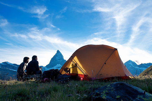 Couple sitting at their orange tent with their Border Collie dog silhouette inside overlooking the Matterhorn mountain at sunset Zermatt Switzerland