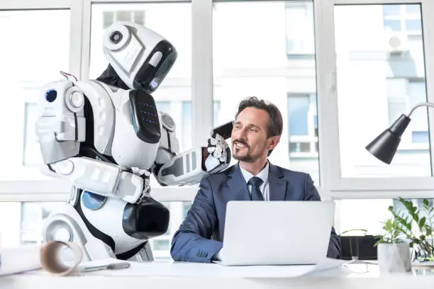 Best helper. Cheerful positive mature man is sitting at table and using laptop while talking on cellphone which is holding big robot