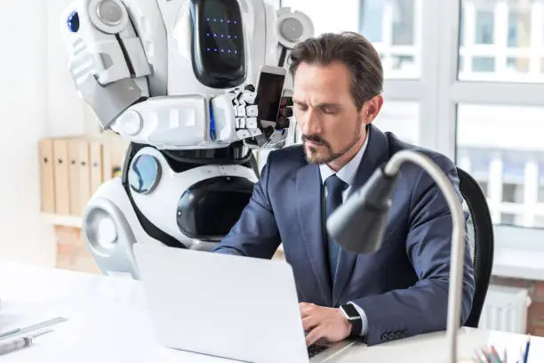 Out of temper. Modern big robot is giving smartphone to cheerless mature businessman who is sitting at table and using laptop