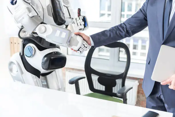 Good work. Close up arms of employee and cyborg. Man in suit is shaking hands with robot against window with cityscape on the background. Droid is showing thumb-up