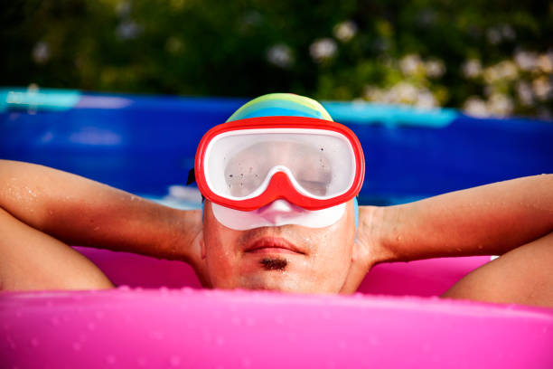 closeup of a young caucasian man wearing a diving mask, a bath cap and a bright pink swim ring relaxing in a portable swimming pool placed in the backyard