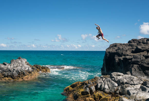 Fit young man jumps into ocean at Lumahai beach Kauai Dangerous leap into warm blue ocean off rocks at Lumahai Beach on Hawaiian island of Kauai north shore stock pictures, royalty-free photos & images