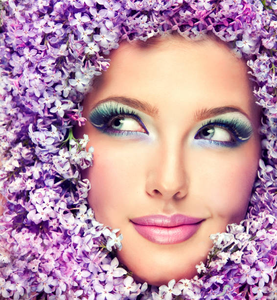 Gorgeous womans face surrounded by lilac flowers.Bright lavender-color make up. Tricky smile with glance to the side. Image and symbol of youth and spring freshness.