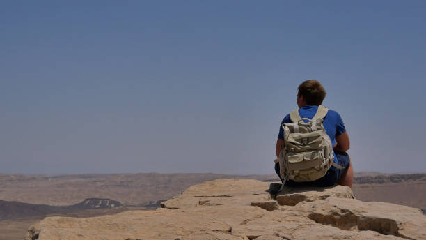istock Young man with backpack sitting on cliff's edge and enjoying the desert view 802673766