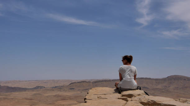 istock Young woman sitting on cliff's edge and looking at the desert 802674080