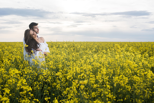 Loving couple embracing while standing in a filed full of yellow flowers and looking away. Copy space.