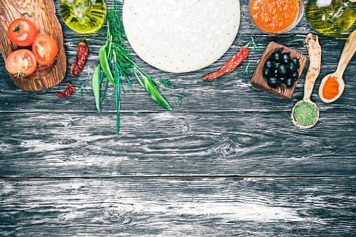 Pizza ingredients - round dough base, fresh tomatoes, sauce, olive oil, green herbs and spices on background of black textured wooden boards. Top view. Copy space