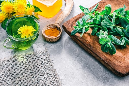 Green mint tea in translucent glass tea cup and honey in rustic spoon on concrete background with dandelions and fresh mint