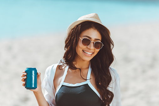 Beautiful young woman in sunglasses and straw hat holding soda can and smiling at camera