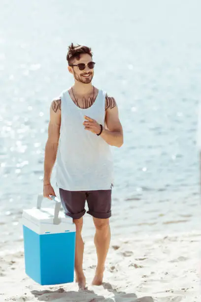smiling young man in sunglasses holding cooler box while standing on beach