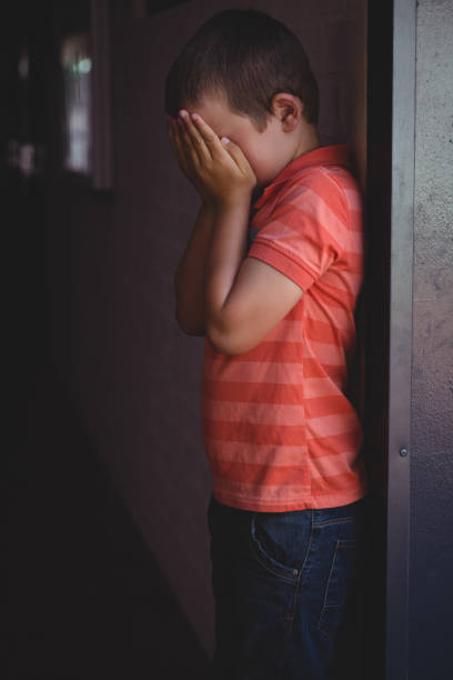 istock Side view of boy covering his face with hands while standing in corridor 803077498