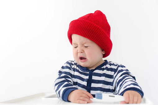 The baby sneezes sitting in a chair in a red hat and a vest with a thermometer in his hand, his eyes are closed