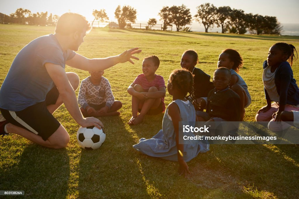 Los niños de primaria y profesor sentado con la bola en campo - Foto de stock de Voluntario libre de derechos