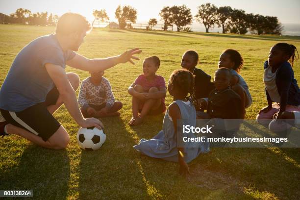 Los Niños De Primaria Y Profesor Sentado Con La Bola En Campo Foto de stock y más banco de imágenes de Voluntario