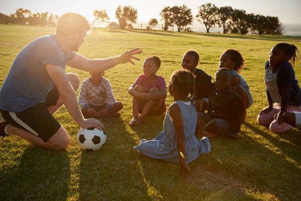 istock Elementary school kids and teacher sitting with ball in field 803138264