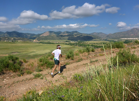 During a summer morning in the grasslands of Bear Creek Lake Park, a young man with a beard runs a trail past the Front Range Rocky Mountains, Morrison, Colorado.
