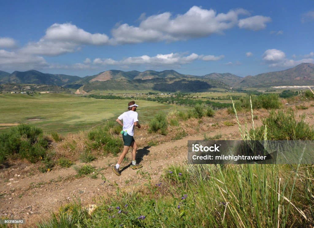Young man trail running Bear Creek Lake Park Front Range Rocky Mountains Colorado During a summer morning in the grasslands of Bear Creek Lake Park, a young man with a beard runs a trail past the Front Range Rocky Mountains, Morrison, Colorado. Front Range - Mountain Range Stock Photo