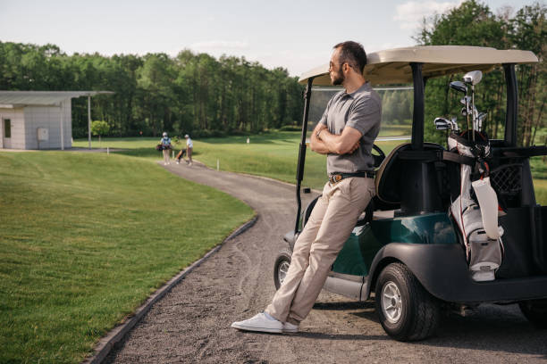ゴルフ場でゴルフのカートの近くに立っている間よそ見物思いにふける男 - golf cart golf bag horizontal outdoors ストックフォトと画像