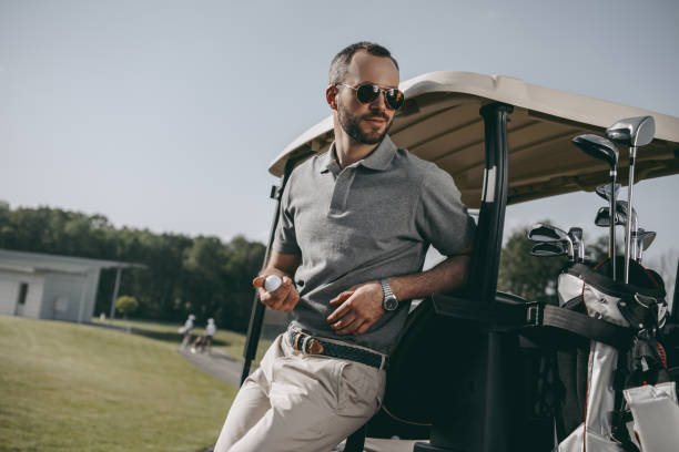 ゴルフ ・ ボールを押し、ゴルフカートにもたれながらよそ見スタイリッシュなゴルファー - golf cart golf bag horizontal outdoors ストックフォトと画像