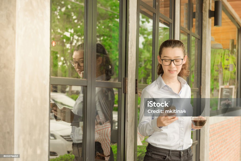 Half Thai half American business woman hold iphone in right hand and glass of hot black coffee in left hand standing against glass door at coffee shop 20-29 Years Stock Photo