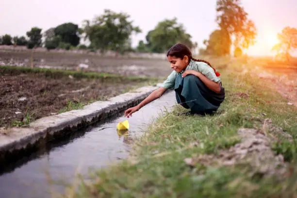 Little girl of elementary age wearing school uniform sitting near water canal outdoor in the field & flowing paper boat in to the water.