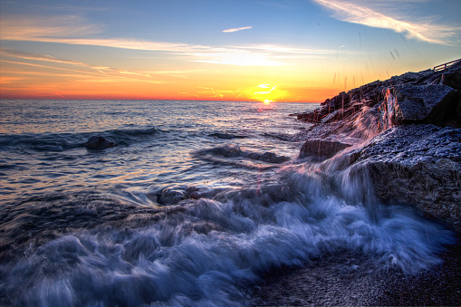 Waves splash on the rocky shore of Lake Huron with a vibrant sunrise at the watery horizon. Lexington, Michigan.