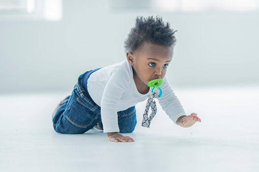 One baby of African descent is crawling inside on white flooring with windows in the background. The baby boy looks curious and is motivated learning to crawl. The child has a green pacifier in his mouth.