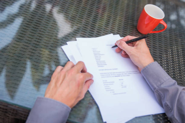 Business men filling questionaire form. His hand writing on top the table. There is a red mug on the table. The man is sitting  outdoor.. He is wearing a gray shirt.