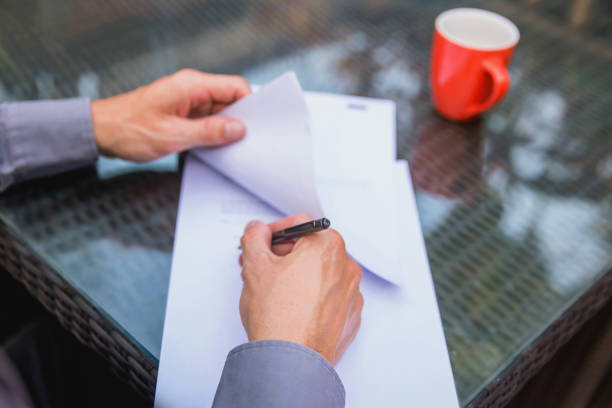 Business men signing legal document.His hand writing on top the table. There is a red mug on the table. The man is sitting  outdoor, in the afternoon. He is wearing a gray shirt.