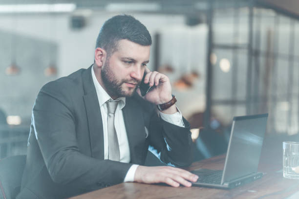 istock Young bearded businessman sitting at table in cafe,holding smartphone and using laptop 803617166
