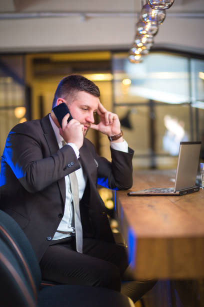istock Young bearded businessman sitting at table in cafe,holding smartphone and using laptop 803617174