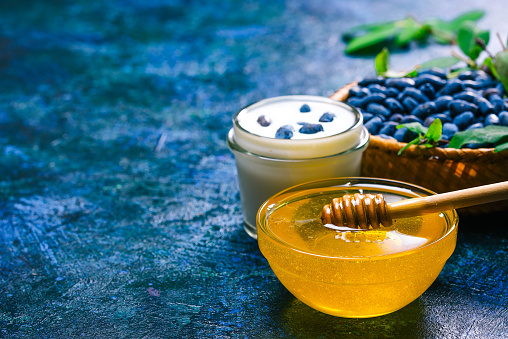 Glass bowl of honey, homemade yogurt and fresh honeysuckle berries in little basket  over spotty blue background. Close up