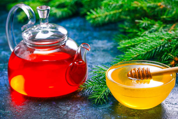 Honey, tea and evergreen twigs Glass bowl of honey, herbal tea in teapot and evergreen fir tree twigs over spotty blue background. Close up pine needle tea stock pictures, royalty-free photos & images