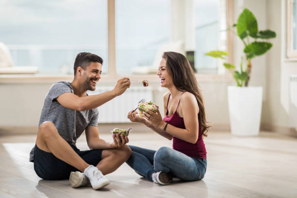 hombre feliz alimentando a su novia mientras estaba sentado en el piso en su ático nuevo. - healthy feeding fotografías e imágenes de stock