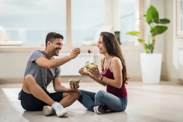 Photo of Happy man feeding his girlfriend while sitting on the floor at their new penthouse.