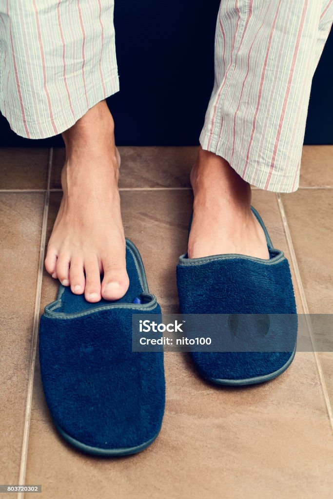 young man putting on or off his slippers closeup of a young caucasian man in pajamas sitting on the edge of the bed putting on or putting off a pair of warm slippers Slipper Stock Photo