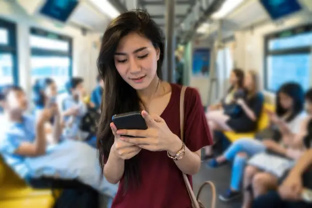 Photo of Asian woman passenger with casual suit using the social network via smart mobile phone in the BTS Skytrain rails or MRT subway for travel in the big city, lifestyle and transportation concept