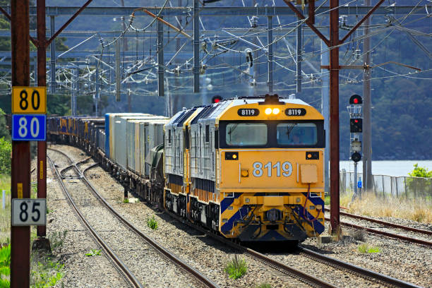 Hawkesbury River, Australia - Oct 28, 2015: Pacific National freight train hauled by two diesel-electric locomotives with heavy load of steel and containers behind, drifts down off the Hawkesbury River Bridge under inter-urban overhead electric wires.