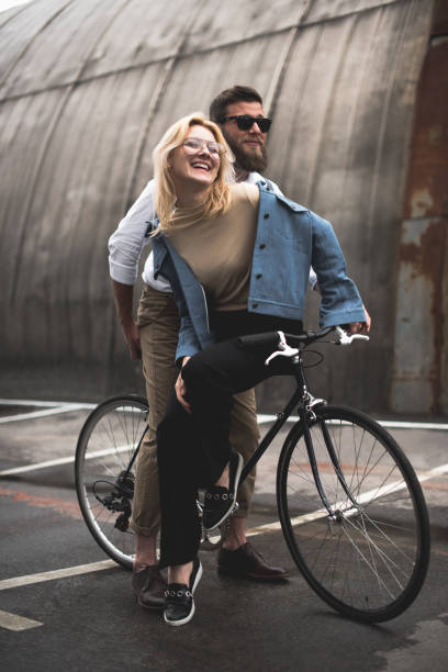 stylish cheerful young couple riding bicycle on parking place - cycling bicycle hipster urban scene imagens e fotografias de stock