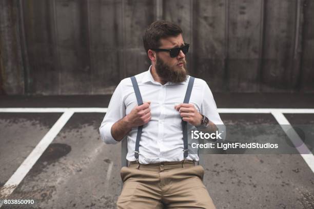 Elegante Uomo Barbuto Con Occhiali Da Sole E Bretelle Seduti Sul Parcheggio E Distospendi - Fotografie stock e altre immagini di Adulto