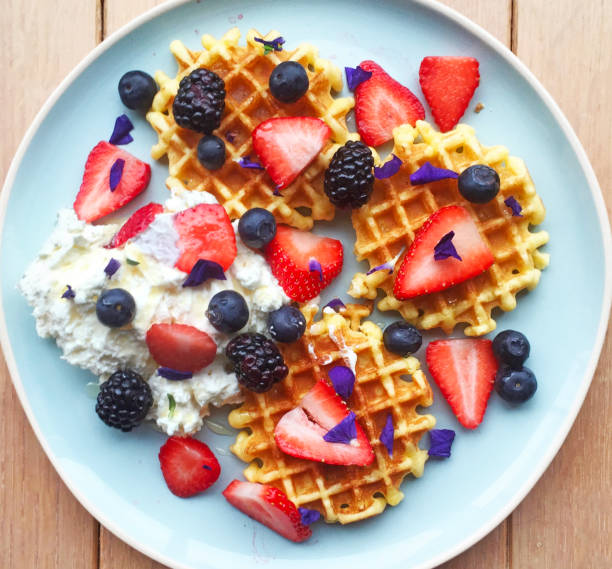 gaufres belges, garnies de crème fouettée, fruits, sirop et fleurs comestibles. vue aérienne de petit déjeuner maison sur fond en bois rustique. - waffle waffled belgian waffle food photos et images de collection