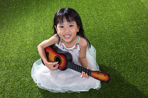 Asian Little Chinese girl sitting on grass and playing with guitar at outdoor park