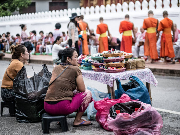 Buddhist monks collecting alms in Luang Pragang Laos while a group of women is selling goods to the tourists so they can give it to the monks. Every morning the monks walk through the streets of Luang Prabang too collect alms of residents and tourists of the town. Although it is recommended to give rice , fruits and vegetables and other fresh food to the monks dozens of vendors wait on the street to sell packaged food to the monks that they throw away as they don't eat processed food. They disposed packaged are picked up by poor people or by the same vendors that resell it again.