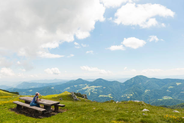 Senior woman hiking in the mountains, she sits on the bench and enjoying the view on mountain range. Sunny and warm.