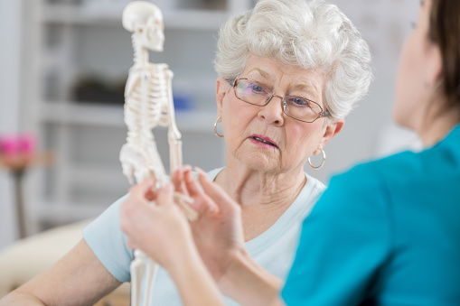 Senior woman listens seriously as her physical therapist sits with her and holds a model of the human skeletal system.  The therapist is discussing the affects of osteoporosis in posture and the reason for resulting pain.
