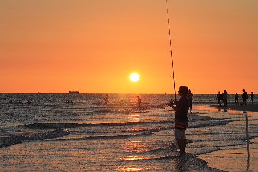 A young man fishes in the Gulf of Mexico at St. Pete Beach, Florida, USA. Other people are in the water or standing on the beach, waiting for sunset. Low sun, dramatic orange sky.