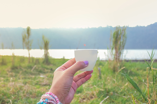 Females hands toasting tea with a grass and lake background, on the camping